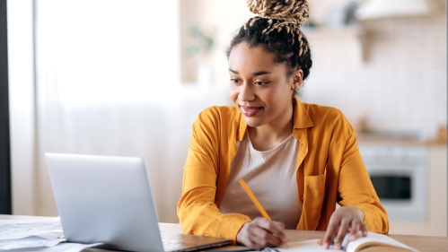 Stock image with young female presenting person sitting at a desk looking at laptop with a yellow shirt on