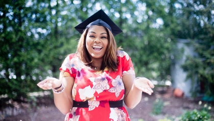 Black female in a red and white floral dress smiling and facing the camera with a graduation cap on