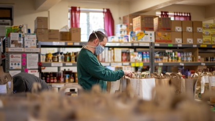 Large room with shelves of food and a person at the center with a medical mask on to cover their mouth and nose while they pack groceries and organize food