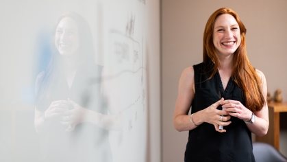 Woman with long red hair, smiling and standing in from of a dry erase board 