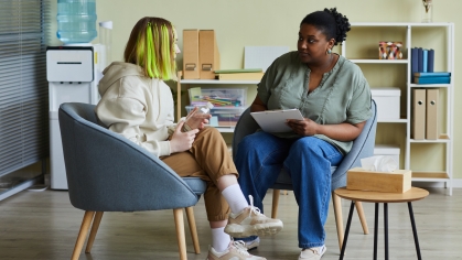 Stock image of two people in a classroom style space. Both are sitting in chairs; one with a notepad and listening as person 2 is talking