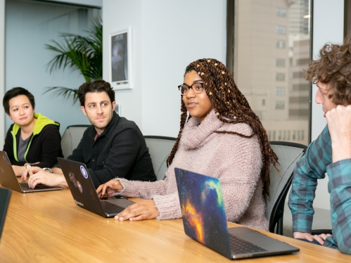 Image of four people sitting at a conference table with their laptops open and talking