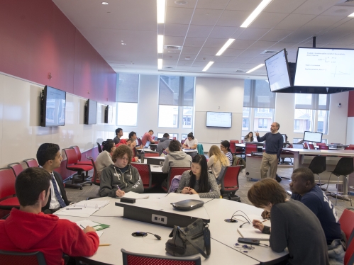 Large classroom with shared tables; students sitting and working with professor lectures at the Center of the room