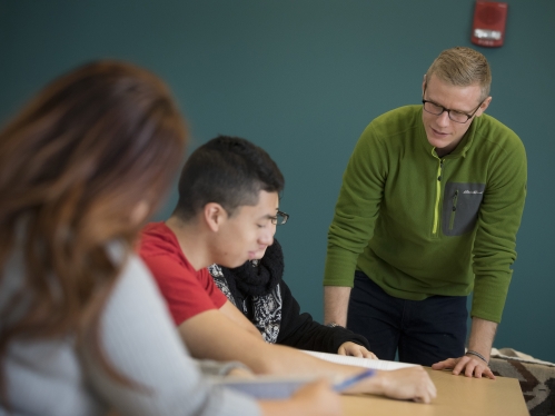 Three people in the frame with one person speaking to the students/people working at a shared table