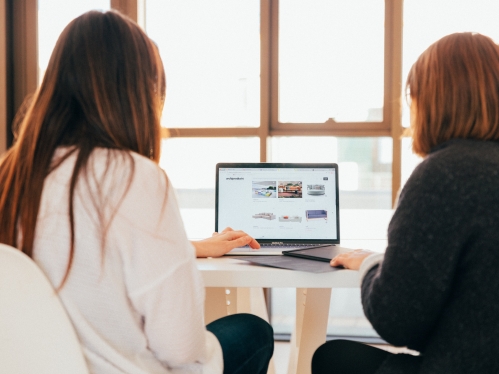 Two female presenting people with their backs to the camera looking at a laptop together