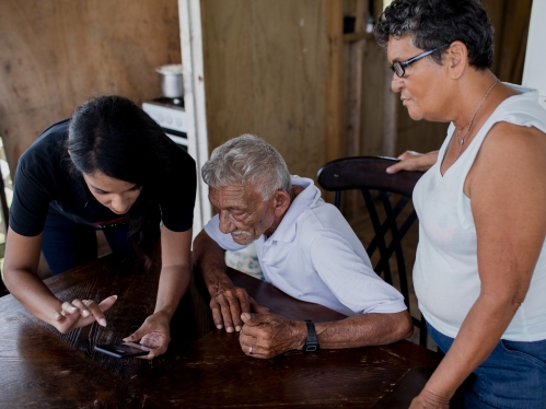 Three people of various ages with one person sitting at a table, they are all looking at a cell phone together