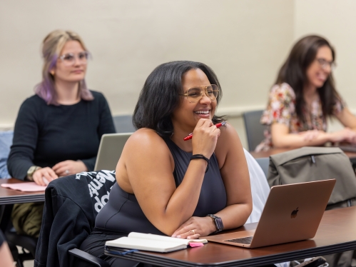 Photo of adult learners in a classroom; focus is on a central female presenting student who is smiling and holding her chin in her hand