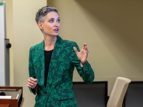 Image of a slender person speaking and looking away from the camera; they are wearing a green patterned suit